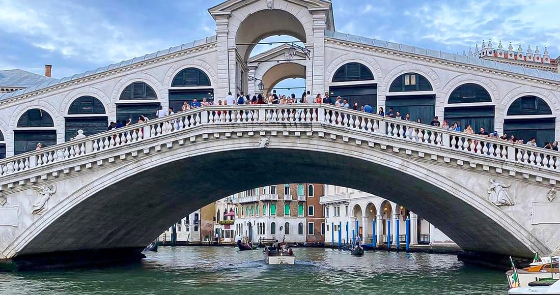 The white-stone Rialto Bridge arching over the Grand Canal in Venice, busy with people, with boats passing beneath