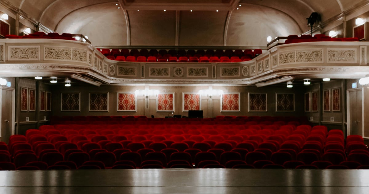 The interior of a grand West End theatre with red seats across the stalls and circle levels