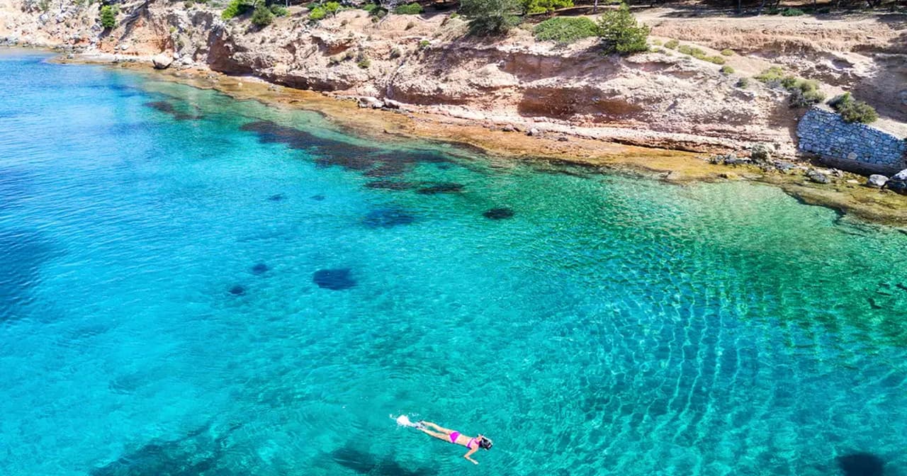 Aerial view of a swimmer in clear turquoise water along a rocky Greek island coastline near Athens
