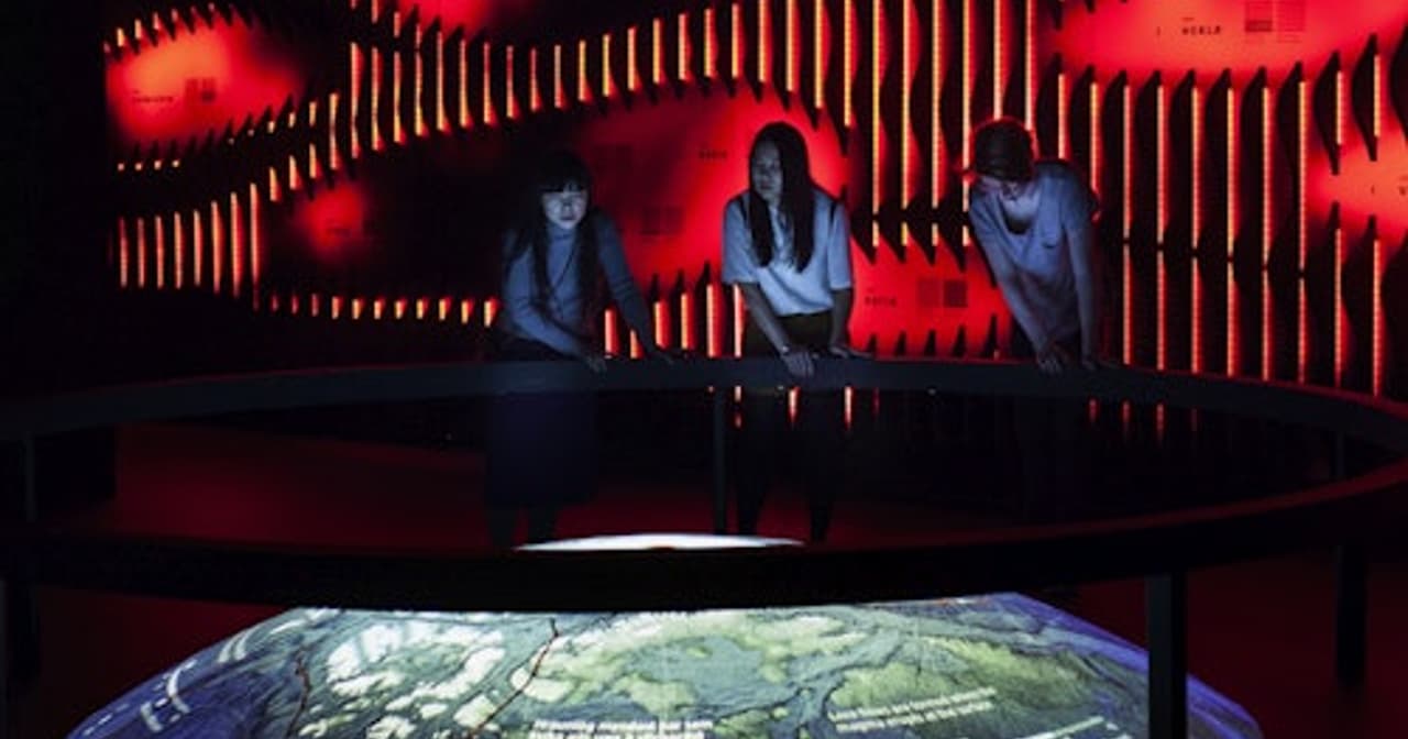 Visitors at a glowing red volcano wall and lit topographic map table in a Hvolsvollur lava exhibition, Iceland