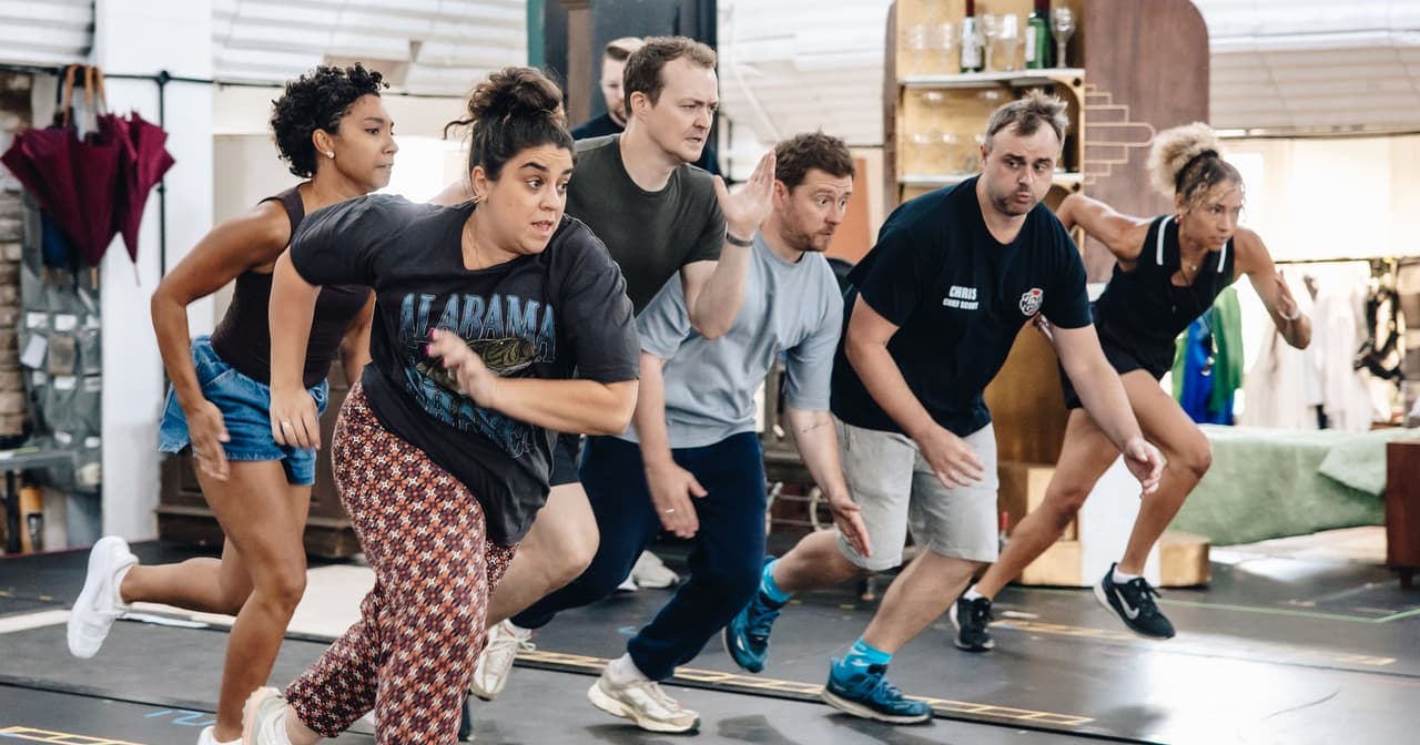 The company of The Comedy About Spies mid-scene in rehearsal, running across the studio floor