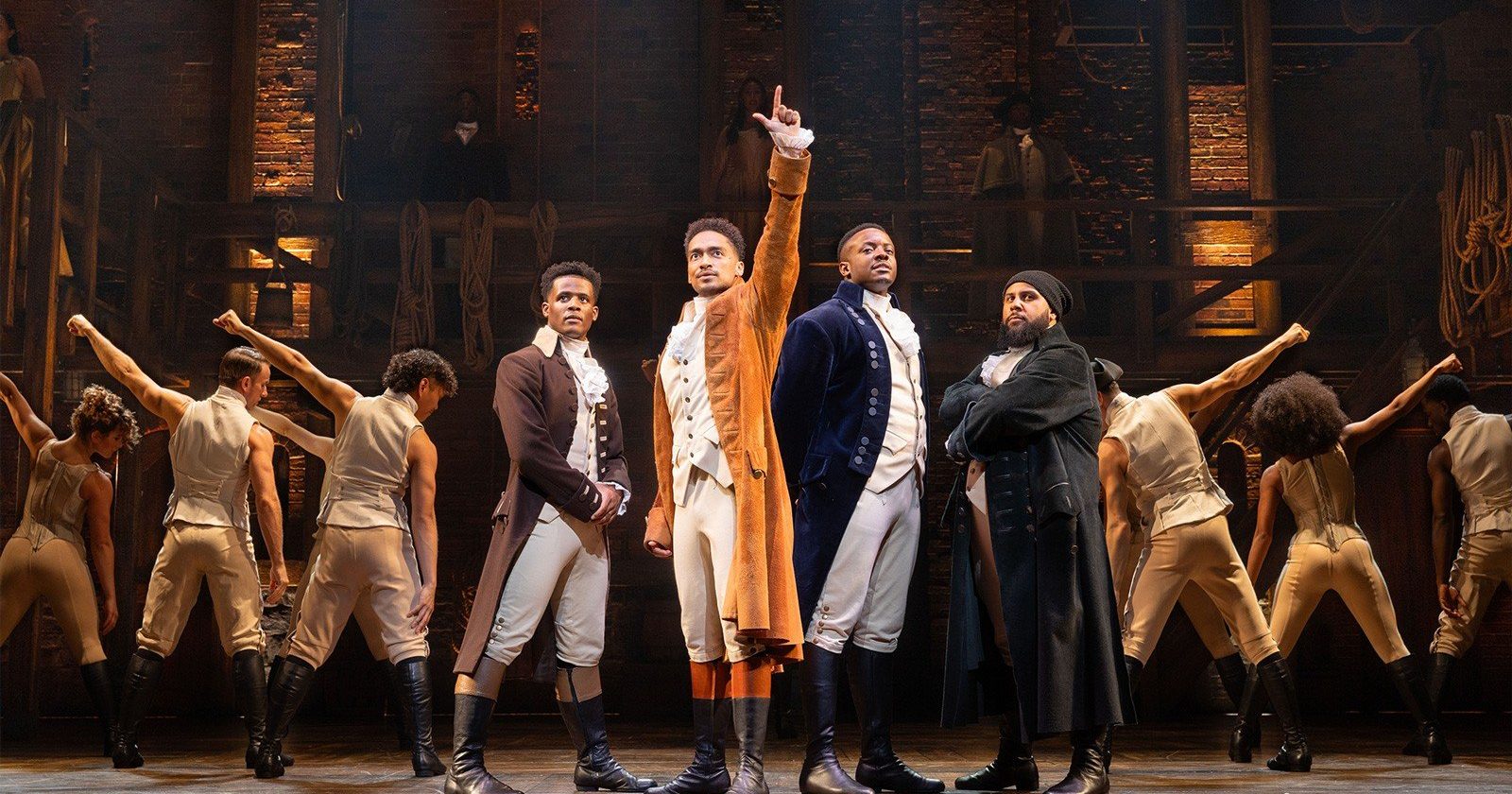 Production image from Hamilton the musical in London. Hamilton stands one arm upstretched, the cast fanning out behind him in a triangle.