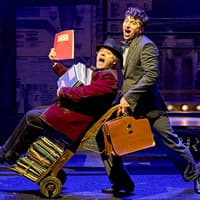 Two actors perform a comedic scene from "The Producers" on stage, one pushing a cart of scripts