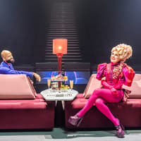 Stage scene from Hunger Games West End show with two actors seated on a pink sofa — The Hunger Games