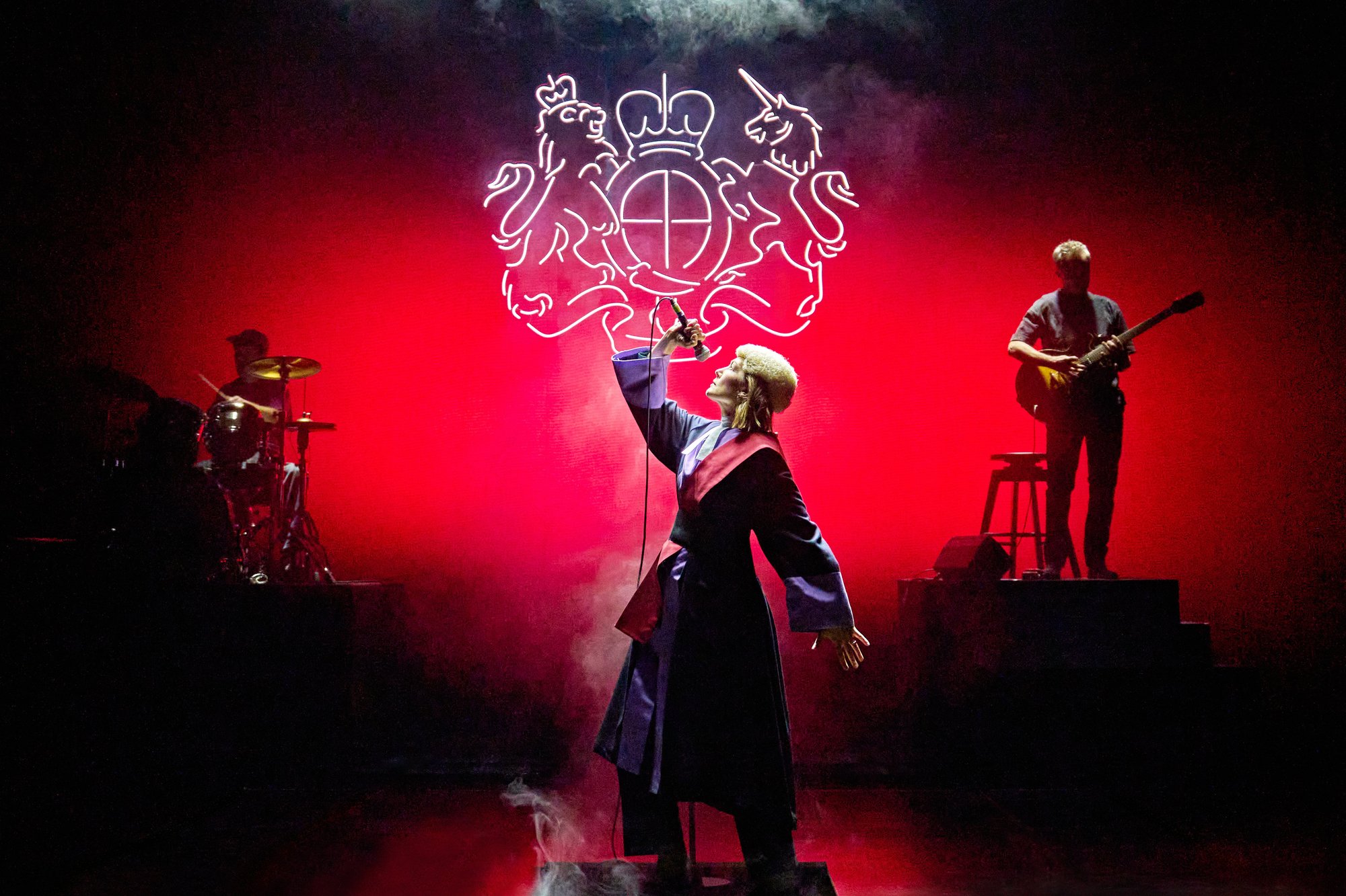 Rosamund Pike in judge’s robes and wig holds a microphone aloft beneath a neon royal coat of arms, with a drummer and guitarist on a red-lit stage in Inter Alia