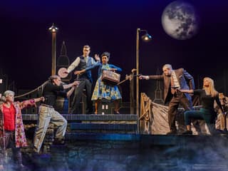 Actors performing a dramatic scene in "The Comedy About Spies" with a cityscape backdrop