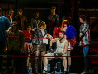 Cast performing a lively scene from Kinky Boots with a boxer in the foreground