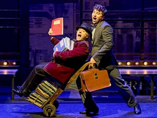 Two actors perform a comedic scene from "The Producers" on stage, one pushing a cart of scripts