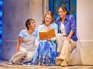 Three actors in a scene from Mamma Mia, sitting on steps with a book, in front of a blue door
