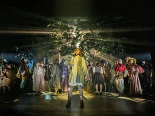 Cast performing in "Into The Woods" with forest backdrop and dramatic lighting