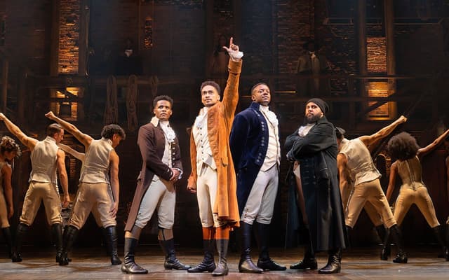 Production image from Hamilton the musical in London. Hamilton stands one arm upstretched, the cast fanning out behind him in a triangle.
