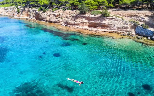 Aerial view of a swimmer in clear turquoise water along a rocky Greek island coastline near Athens