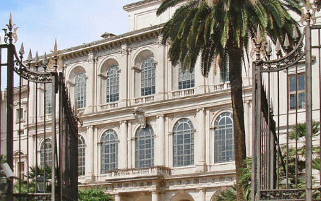 Baroque facade of Palazzo Barberini seen through wrought-iron gates with a palm tree in Rome