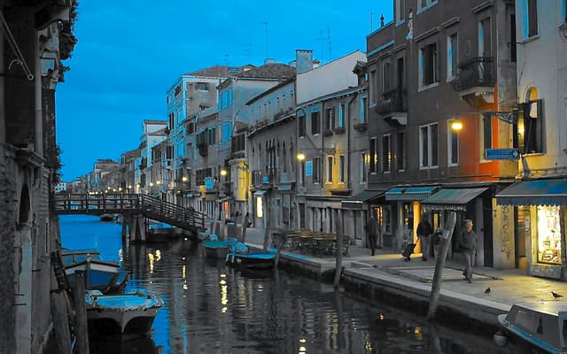 Quiet Venice canal at blue hour with a wooden footbridge, moored boats and lit shopfronts