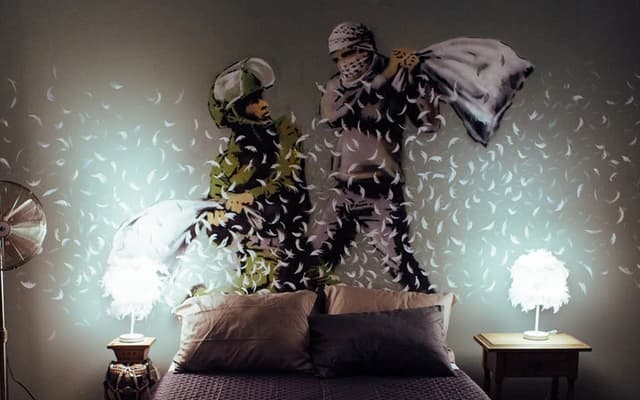 Banksy-style pillow-fight mural of two figures and feathers above a bed in a Barcelona art space