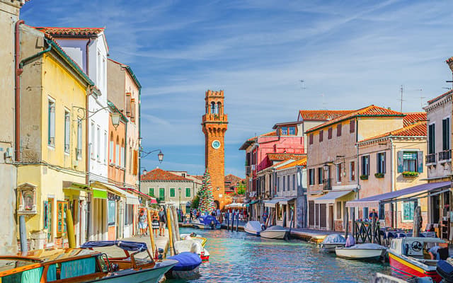 Colourful Murano houses and clock tower along a Venice canal lined with moored boats
