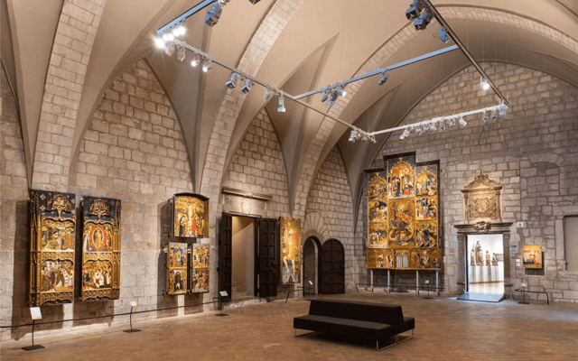 Vaulted stone hall of a Girona museum displaying medieval gold altarpieces under Gothic arches