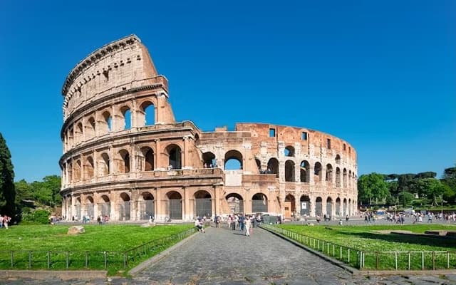 The Colosseum in Rome under a clear blue sky with a path and green lawns in the foreground