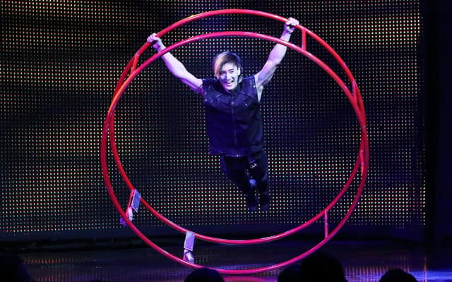 An acrobat spinning inside a large red wheel on stage in V The Ultimate Variety Show
