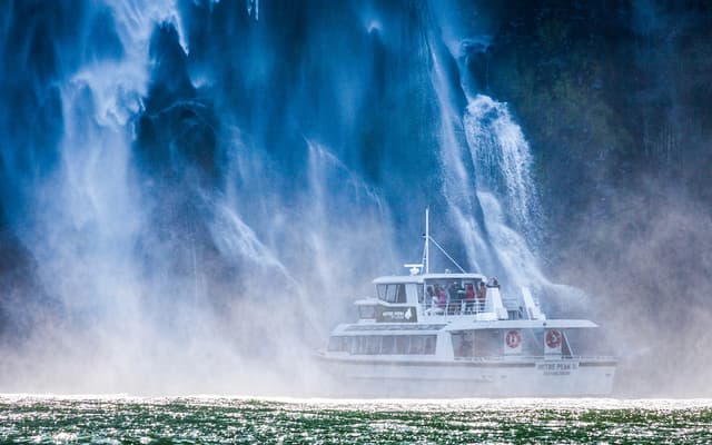 Small cruise boat sailing beneath the sheer cliffs and waterfalls of Milford Sound