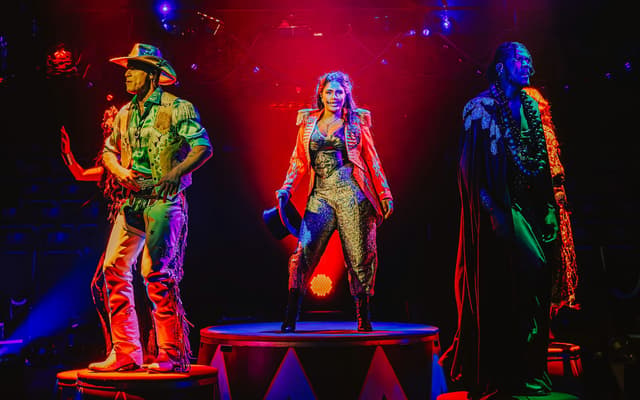 Three costumed performers, including a ringmaster, strike a pose on circus podiums under vivid stage lighting in Come Alive!