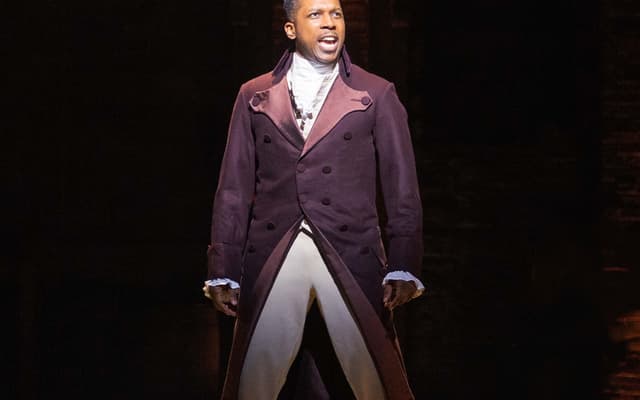 Leslie Odom, Jr. as Aaron Burr, spotlit in a plum coat on stage in Hamilton at the Victoria Palace Theatre