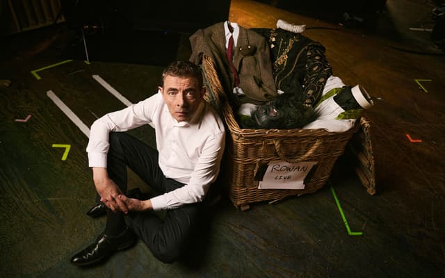 Rowan Atkinson sitting on a theatre stage beside a wicker costume basket labelled Rowan Live