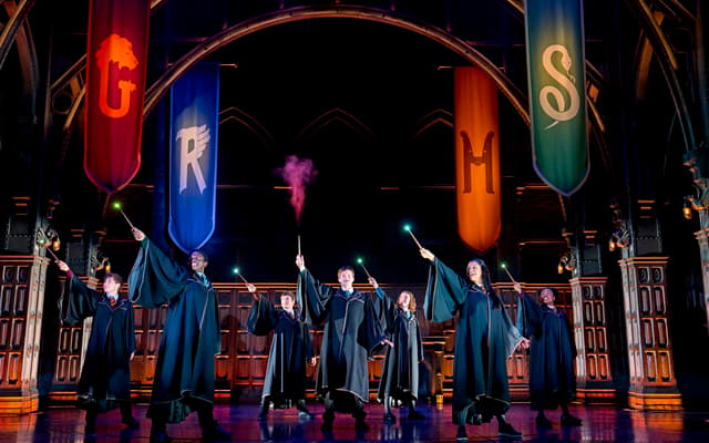 The company of Harry Potter and the Cursed Child raising lit wands beneath the Hogwarts house banners at the Palace Theatre