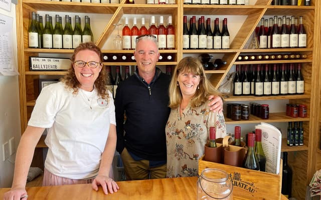 Three smiling people behind the counter of a wine cellar door with shelves of bottles
