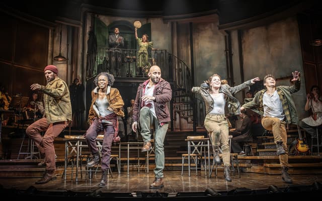 Hadestown cast performing on a tiered wooden stage in worn workwear in the London production