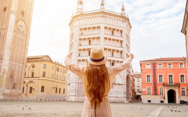 Woman in a sun hat raising her arms before the Parma Baptistery and cathedral tower in Italy