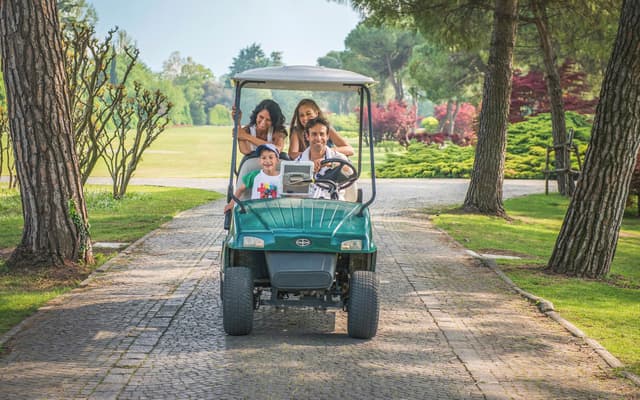 Family of four riding a green golf cart along a tree-lined park path near Verona