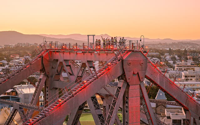 Climbers on the Brisbane Story Bridge summit lit red against a golden sunrise sky