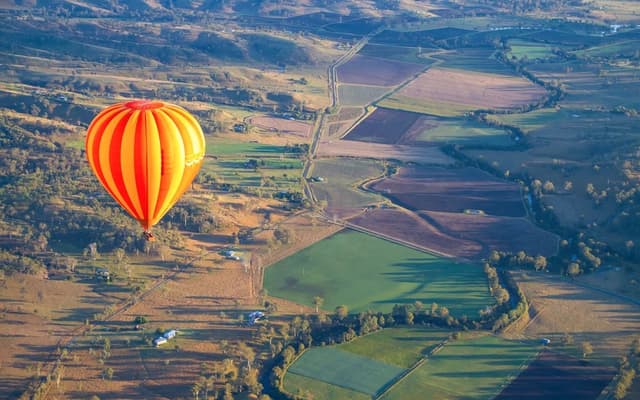 Orange hot air balloon drifting high above patchwork green farmland near Brisbane