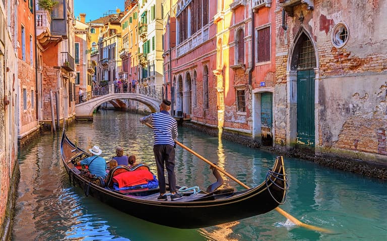 A traditional Venetian gondola with a striped-shirt gondolier gliding along a quiet canal between historic buildings in Venice