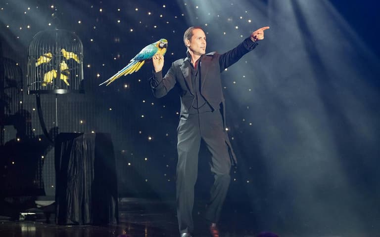 A magician with a blue and gold macaw on stage in V The Ultimate Variety Show
