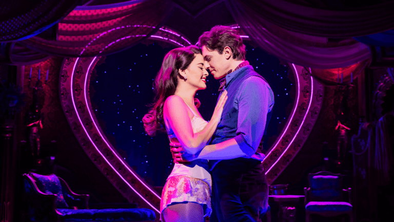 Los protagonistas de Moulin Rouge! se abrazan frente a un corazón de neón luminoso en el escenario del Al Hirschfeld
