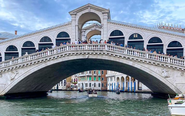 The white-stone Rialto Bridge arching over the Grand Canal in Venice, busy with people, with boats passing beneath