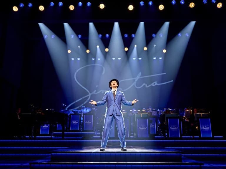 Joel Harper-Jackson como Frank Sinatra, con los brazos abiertos en plena canción bajo reflectores azules en Sinatra: The Musical
