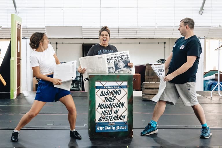 Three cast members brandish newspapers around a Daily News stand reading Diamond Thief Monaghan Held in Amsterdam in rehearsal for The Comedy About Spies
