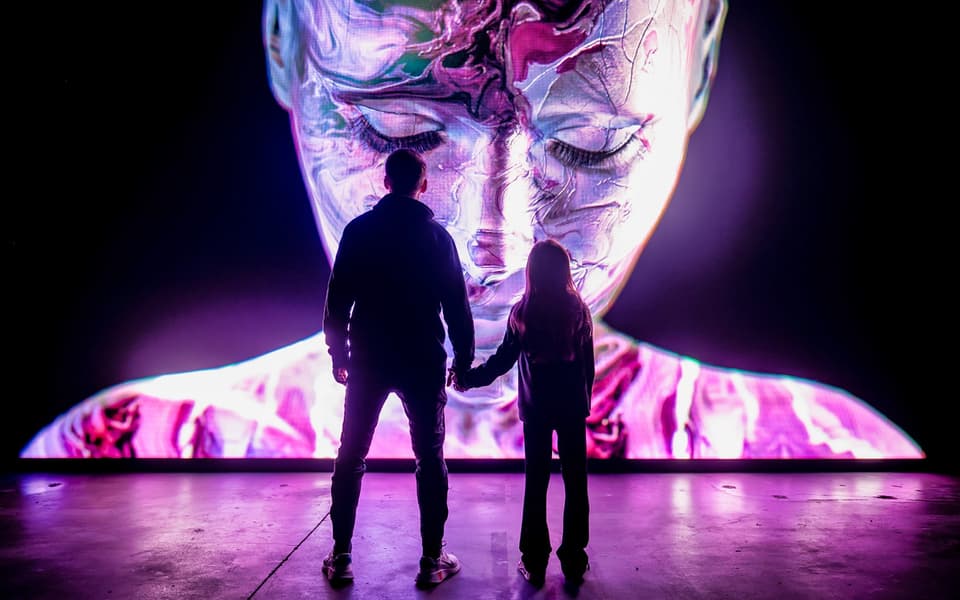Adult and child silhouetted before a giant glowing immersive digital art face in Amsterdam