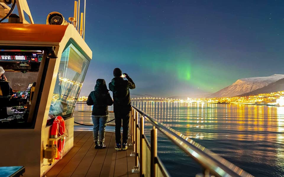 Two people on a boat deck watching the green Northern Lights over the lit-up Tromso fjord at night