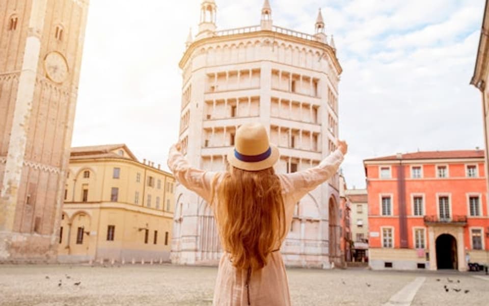 Woman in a sun hat raising her arms before the Parma Baptistery and cathedral tower in Italy