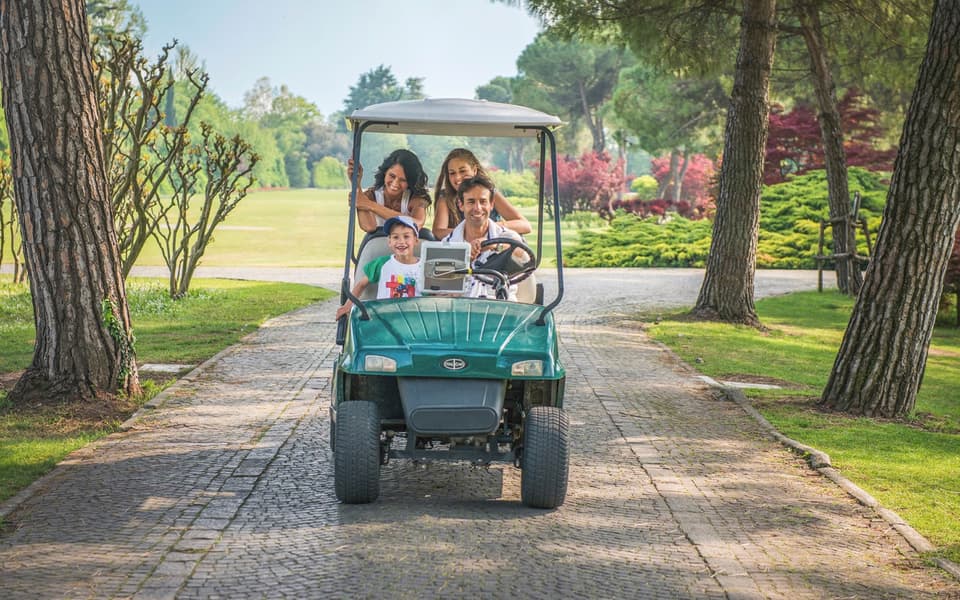 Family of four riding a green golf cart along a tree-lined park path near Verona