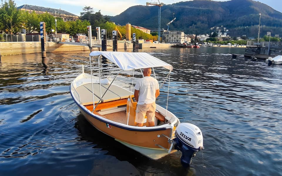 Man steering a small canopied motorboat on Lake Como with the lakeside town and mountains behind