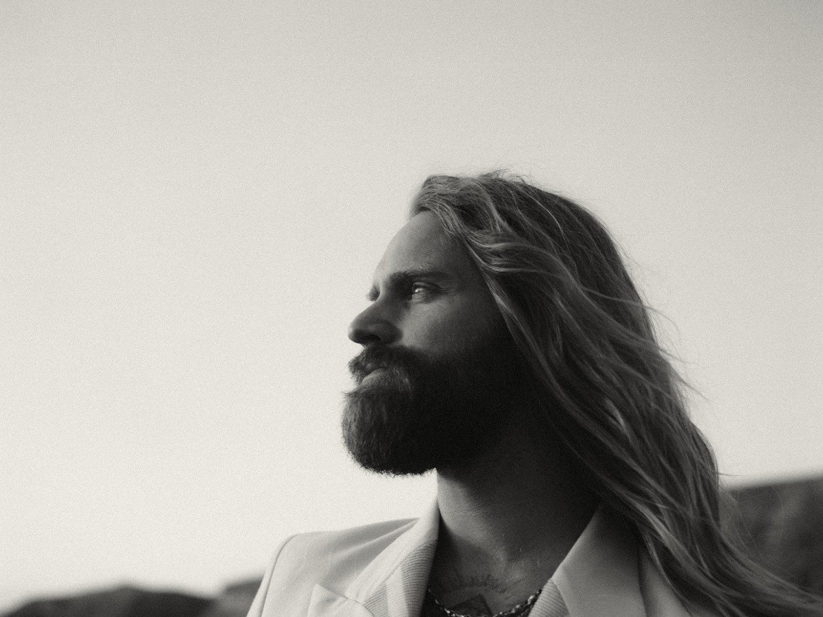 Black and white portrait of Sam Ryder in profile, with long hair and a full beard, wearing a pale jacket